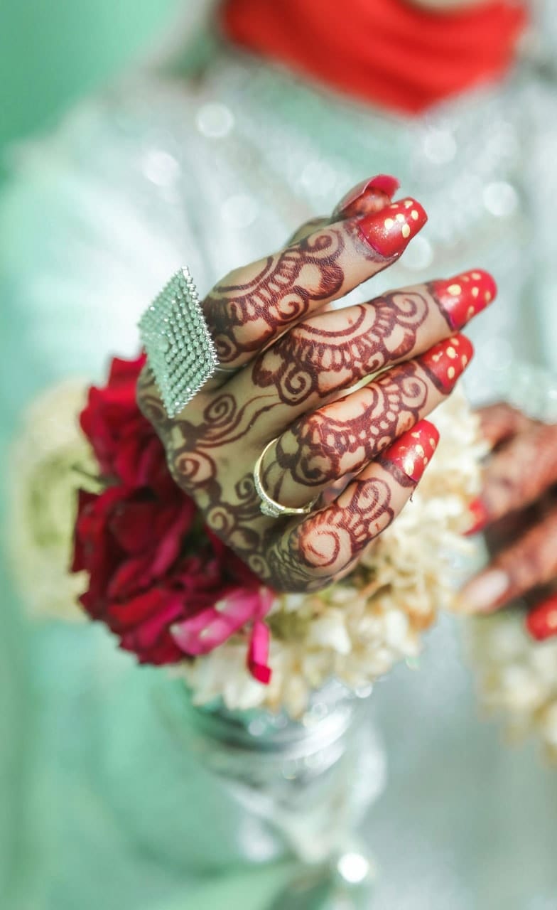 Henna-patterned hand with red nails, wearing a square cocktail ring set with white stones, holding red roses and white flowers